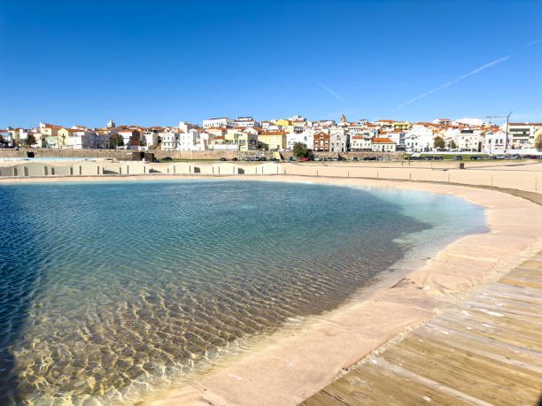 A picturesque view of Figueira da Foz beach in Portugal, featuring a beach pool formed by the ocean waves, with the town's colorful buildings in the background and a clear blue sky.